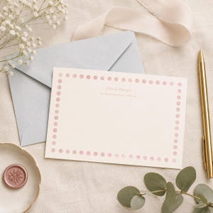 Può includere: Un biglietto rettangolare color crema con un bordo di pois rosa tenue. Il testo Olivia Harper e 111 Buttercup Lane, ABC 123 è stampato al centro. Il biglietto è accompagnato da una busta azzurra e da una penna color oro.