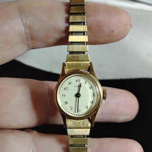 Könnte beinhalten: Eine goldfarbene Timex-Armbanduhr mit einem runden, cremefarbenen analogen Zifferblatt und schwarzen arabischen Ziffern von 1 bis 12. Die Uhr verfügt über ein passendes, dehnbares Metallarmband mit rechteckigen Gliedern und eine kleine Krone.
