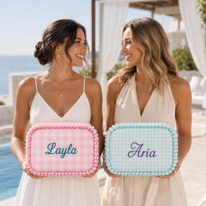 May include: Two rectangular storage cases with a gingham pattern and beaded trim. One case is pink and white with the name Layla embroidered in teal. The other case is light blue and white with the name Aria embroidered in purple script.