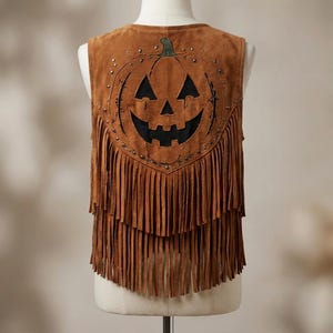 Puede incluir: Un chaleco de ante marrón que presenta un gran diseño recortado de calabaza jack-o'-lantern en la espalda. La prenda está adornada con pequeños tachones metálicos alrededor del motivo de calabaza e incluye dos niveles de flecos largos.