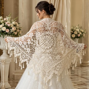 Könnte beinhalten: Ein cremefarbenes, dreieckiges Häkeltuch mit einem komplexen zentralen Mandalamuster, floralen Motiven und einem zarten Netzdesign. Die Kanten sind mit einer Bogenkante und kleinen hängenden Quasten am unteren Rand versehen.