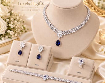 Op de afbeelding: Een sieradenset bestaande uit een ketting, een paar oorbellen, een ring en een armband, bezet met heldere marquise-geslepen stenen en een diepblauwe druppelvormige steen. De ketting toont de tekst Luxebellagifts.