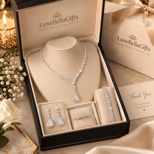 May include: A silver-toned jewellery set in a black LuxeBellaGifts box, featuring a V-shaped necklace with a pear-cut pendant, two drop earrings with pear-cut stones, a tennis bracelet, and a band ring, all adorned with clear, sparkling crystals.