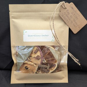 May include: A brown paper pouch with a clear window containing dried orange slices, bay leaves, and spices. A white label reads Guardians Ember. A brown cardboard tag attached with twine is handwritten with the text Guardians Ember Best for Protection grounding and stability.