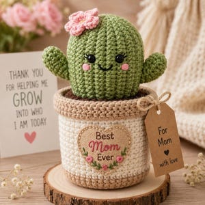 Könnte beinhalten: Ein gehäkelter grüner Kaktus mit einer rosa Blume, schwarzen Augen und einem lächelnden Mund in einem beige-weißen Topf. Der Topf hat einen herzförmigen Aufnäher mit dem Text Best Mom Ever und ein braunes Etikett mit For Mom with love.