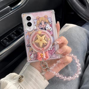 以下が含まれることがあります： セーラームーンとハローキティのグラフィックプリントが施された透明な折りたたみ式スマホケース。ケースにはピンクのリボンが付いた円形の星型ポップアウトグリップと、半透明のピンクのビーズと金属製のチャームで作られたぶら下がり式のリストストラップが付いています。