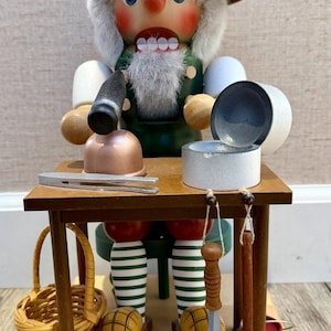Könnte beinhalten: Eine hölzerne Nussknacker-Figur mit beigefarbener Baskenmütze, grauem Haar und grüner Weste, die an einem braunen Tisch sitzt. Auf dem Tisch stehen eine kupferfarbene Schale, ein silbernes Werkzeug und ein weißer Behälter. Korb und Werkzeuge hängen.