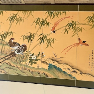 以下が含まれることがあります： 金色の背景に絵が描かれた4枚折りの屏風。竹や岩の近くに止まったり飛んだりしている長い尾の鳥が描かれており、白い花と右側には黒い書道の文字が添えられています。