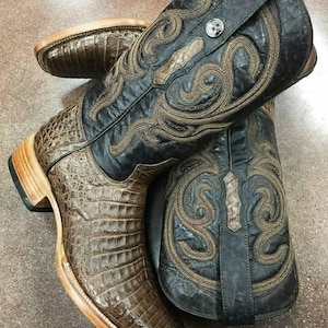 Op de afbeelding: Een paar cowboylaarzen met een bruine, getextureerde voet met een vierkante neus en een zwarte leren schacht. De schacht is versierd met ingewikkeld goudkleurig borduurwerk en een kleine zilverkleurige metalen stud. De hak en zool zijn lichtbruin.