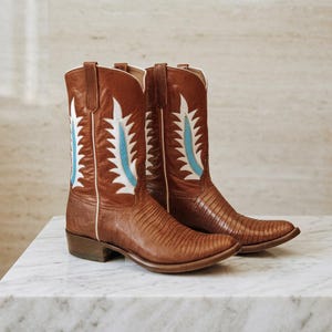 Könnte beinhalten: Ein Paar braune Leder-Cowboystiefel mit einer strukturierten Eidechsenhaut-Optik am Fuß und Schaft. Die Stiefel sind mit weißen und hellblauen, flammenförmigen Lederapplikationen an den Seiten verziert und haben einen Blockabsatz.