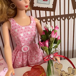 May include: A doll in a pink dress with a white polka dot pattern, holding a bouquet of pink flowers. A heart-shaped Godiva box and a wooden "LOVE" sign are on the table. The doll has blonde hair and gold hoop earrings.