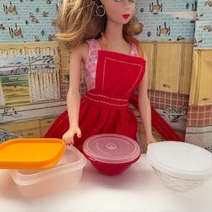 May include: A doll wearing a red apron and dress stands in front of a kitchen backdrop. Several plastic food containers with lids are in the foreground. One container is orange, one is clear, and one is red.
