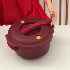 May include: A miniature burgundy-colored pressure cooker with a matching lid and handle. The lid features two yellow circular accents and a red handle. The cooker is designed for a doll or toy set.