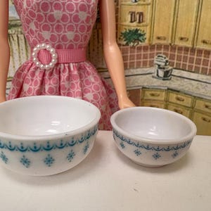 May include: Two white ceramic bowls with a blue floral pattern. The bowls are different sizes, with the larger bowl on the left and the smaller bowl on the right. The bowls are set against a background of a doll wearing a pink dress.