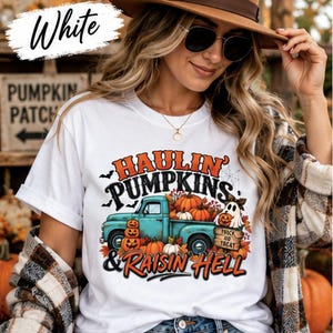 May include: A white t-shirt featuring a graphic of a teal pickup truck loaded with orange pumpkins and a ghost. The text reads Haulin' Pumpkins & Raisin Hell, with Trick or Treat on a wooden sign. The design includes autumn leaves and bats.