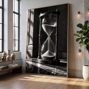 May include: A large rectangular wall art print featuring a black and white photograph of a classic hourglass with dark metal framing. The hourglass is positioned on a textured surface, with sand flowing from the top bulb into the bottom bulb.