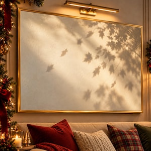 May include: A large rectangular wall mirror with a thin gold frame, reflecting a soft, warm light and the distinct shadows of maple leaves cast across its surface. A horizontal gold light fixture is mounted directly above the mirror frame.