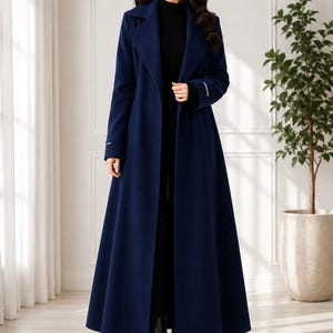 May include: A floor-length navy blue wool-blend coat featuring a wide notched lapel, long sleeves with white cuff detailing, and a structured silhouette. The coat is worn over a black ribbed turtleneck jumper and black leather ankle boots.
