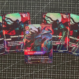 Könnte beinhalten: Fünf rechteckige Sammelkarten mit Dark-Fantasy-Illustrationen. Vier Karten sind mit Shadow Clone beschriftet, eine zentrale Karte mit Master of Shadows. Die Karten zeigen rote, blaue und schwarze Farben sowie Text zu Spielmechaniken.