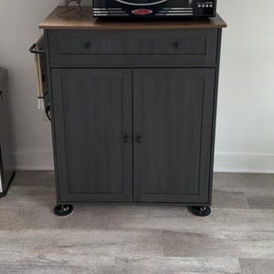 May include: A dark grey kitchen trolley with a wood-effect top, one drawer, and two cabinet doors on castors. A black Nostalgia Electrics microwave sits on top next to a small speckled rooster figurine. A towel hangs from a side bar on the left.