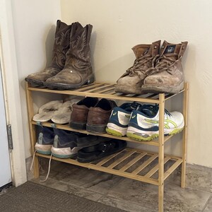 Pode incluir: Um sapateiro de madeira clara de três níveis contendo vários calçados, incluindo botas de cowboy de couro marrom, botas de trabalho bege, tênis esportivos brancos e azuis, mocassins marrons, chinelos brancos felpudos e sandálias pretas.