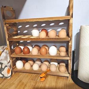 May include: A three-tiered wooden egg rack with a scorched finish, holding numerous brown and white chicken eggs. The rack features circular cutouts on the side panels and is positioned next to a roll of white paper towels and a patterned cloth.