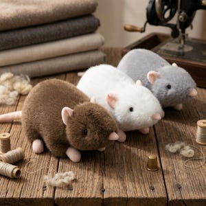 Puede incluir: Tres juguetes de peluche con forma de rata en marrón, blanco y gris, con pelaje suave, orejas pequeñas rosadas y nariz puntiaguda rosada. Los roedores están sobre una superficie de madera junto a carretes de hilo y un dedal.