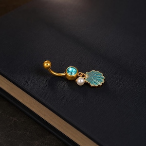 Può includere: Un piercing per l'ombelico curvo color oro con un cristallo rotondo blu brillante sulla parte superiore, una piccola perla bianca e un ciondolo a forma di conchiglia tempestato di minuscole pietre blu scintillanti.