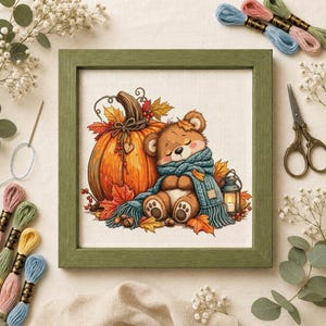 Peut inclure: Une broderie au point de croix encadrée représentant un ours en peluche marron portant une écharpe bleu sarcelle, assis à côté d'une grosse citrouille orange et d'une petite lanterne, le tout décoré de feuilles d'automne dans un cadre en bois vert.