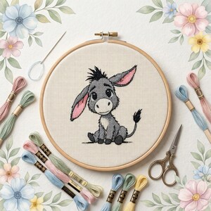 May include: A cross-stitch project in a round wooden hoop featuring a gray donkey with large ears and a tufted tail. The kit includes a silver needle, vintage-style metal scissors, and several skeins of embroidery floss in pastel shades.