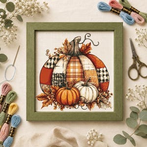 May include: A framed cross-stitch embroidery featuring a large central pumpkin with a patchwork design of orange, white, and black plaid patterns, accompanied by two smaller pumpkins and autumn leaves, surrounded by embroidery floss and scissors.