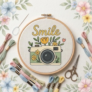 Könnte beinhalten: Ein Stickrahmen mit einem Stoffeinsatz, der eine gestickte Illustration einer Vintage-Kamera, gelbe Blumen und das Wort Smile zeigt. Das Set enthält eine Nadel, Stickgarn in verschiedenen Pastellfarben und eine Metallschere.