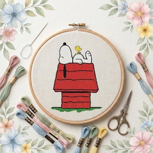 May include: A cross-stitch embroidery project in a wooden hoop featuring Snoopy and Woodstock on a red doghouse. The design is centered on white fabric, surrounded by colorful embroidery floss skeins, a needle, and vintage-style scissors.