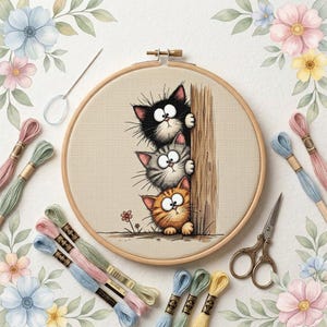 May include: A circular wooden embroidery hoop containing a cross-stitch pattern of three stacked cats—one black, one grey tabby, and one orange tabby—peeking from behind a wooden post, surrounded by embroidery floss, a needle, and scissors.