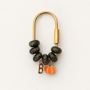 May include: A gold-toned metal carabiner keychain featuring five dark, translucent, rounded beads threaded onto the frame. Two small charms hang from the bottom: a gold-colored letter B and an orange enamel pumpkin with a green stem detail.