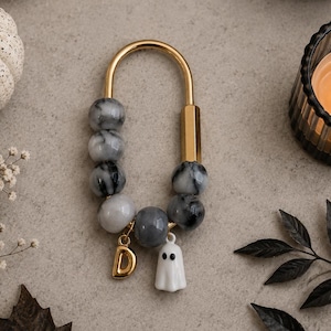 May include: A gold-toned U-shaped metal keychain featuring a series of grey and white marbled beads. The keychain is accented with a small gold-colored letter D charm and a miniature white ghost charm with two black eyes hanging from the beads.
