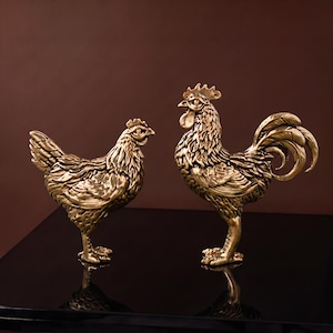 Op de afbeelding: Twee metalen goudkleurige beelden van een kip en een haan. Beide figuren hebben gedetailleerd, gestructureerd verenkleed, gedefinieerde kammen en lellen. De haan staat rechtop met een prominente staart, terwijl de kip een lager profiel heeft.
