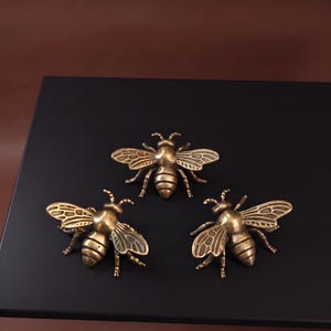 Op de afbeelding: Drie identieke bijvormige decoratieve ornamenten van messing met gedetailleerde vleugels met geëtste lijnen, een afgerond borststuk en een gesegmenteerd achterlijf. Elk metalen insectfiguur heeft een gepolijste afwerking.