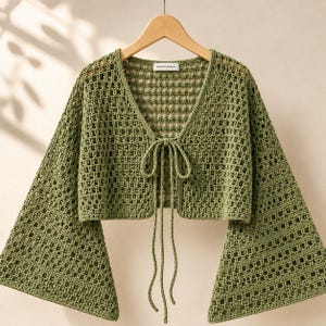 May include: An olive green crochet bolero featuring an open-knit square pattern, a V-neckline, and long flared sleeves. The garment includes a front tie closure with two long matching strings and a small white label at the inner neckline.