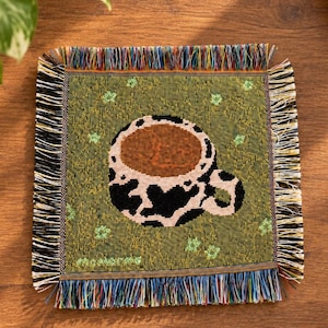 May include: A square woven coaster featuring a green background with small light green star patterns and a central graphic of a coffee mug with a black and white cow print design, finished with multicoloured fringed edges on all four sides.