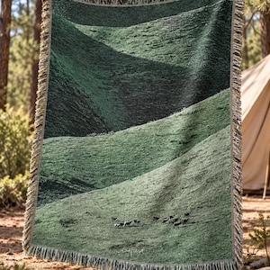 May include: A rectangular woven throw blanket featuring a dark green landscape design of rolling hills and a small herd of grazing animals. The blanket has a textured surface and a decorative fringed edge along all four sides of the fabric.