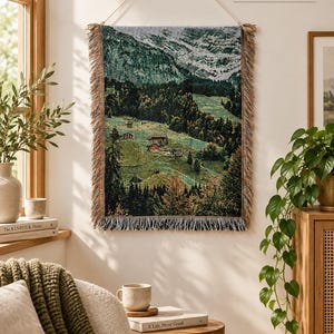 Könnte beinhalten: Ein rechteckiger, gewebter Wandteppich mit einer Landschaft aus grünen Bergen, dichten Wäldern und kleinen Häusern. Der Wandteppich hat an den Seiten und am unteren Rand Fransen, hängt an einer Schnur und misst laut Aufdruck 60 x 80 cm.