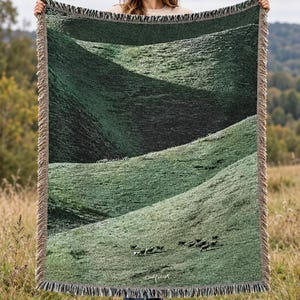 May include: A rectangular woven throw blanket featuring a dark green landscape of rolling hills and a small herd of sheep grazing in the foreground. The blanket has a fringed border on all four sides and includes a small white signature at the bottom.