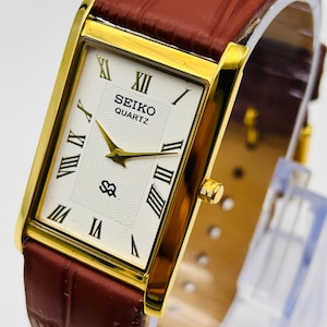 Puede incluir: Un reloj de pulsera rectangular Seiko Quartz con caja dorada y esfera blanca con números romanos negros. El reloj presenta un patrón central texturizado, manecillas doradas y una correa de cuero marrón con textura de piel de cocodrilo.