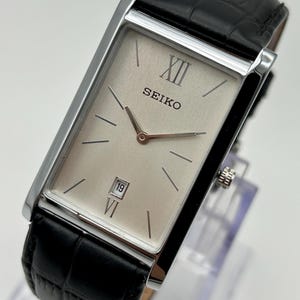 Op de afbeelding: Een rechthoekig Seiko-polshorloge met een zilverkleurige wijzerplaat, zwarte Romeinse cijfers op de twaalf- en zesuurpositie en dunne zilveren streepindexen. Het horloge heeft een datumvenster op de zesuurpositie en een zwarte lederen band.