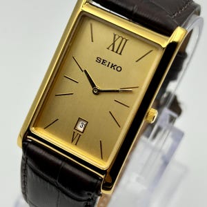 May include: A rectangular Seiko wristwatch with a gold-tone dial, gold hands, and gold hour markers. The watch features Roman numerals at the twelve and six o'clock positions, a date window at six, and a dark brown embossed leather strap.