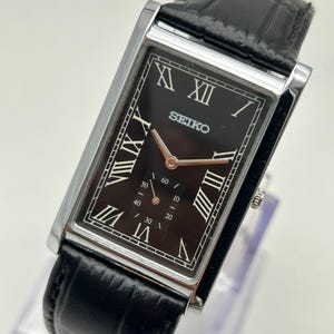 May include: A rectangular Seiko wristwatch with a black dial, silver-tone case, and black leather strap. The dial features white Roman numeral hour markers, rose gold-tone hands, and a small sub-dial at the bottom with numerical markings.