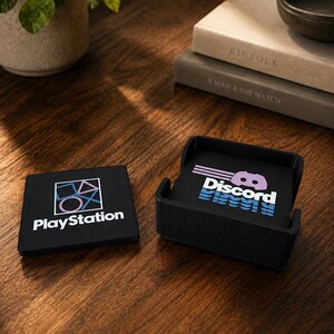 May include: A set of square black coasters featuring the PlayStation logo with its iconic geometric shapes and the Discord logo with its stylized controller icon and text. The coasters are stored in a black ribbed holder with one coaster placed outside.