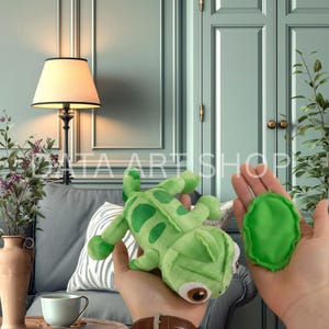 Può includere: Un peluche a forma di camaleonte verde con macchie verde scuro e grandi occhi marroni, accompagnato da un piccolo elemento ovale in tessuto verde. Il giocattolo presenta una consistenza morbida e un design dettagliato, tenuto in mano.