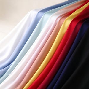May include: A collection of smooth, draped fabric swatches arranged in a gradient of colors including white, light blue, pale pink, yellow, red, dark blue, and black, showcasing a soft texture and flowing folds across the surface.